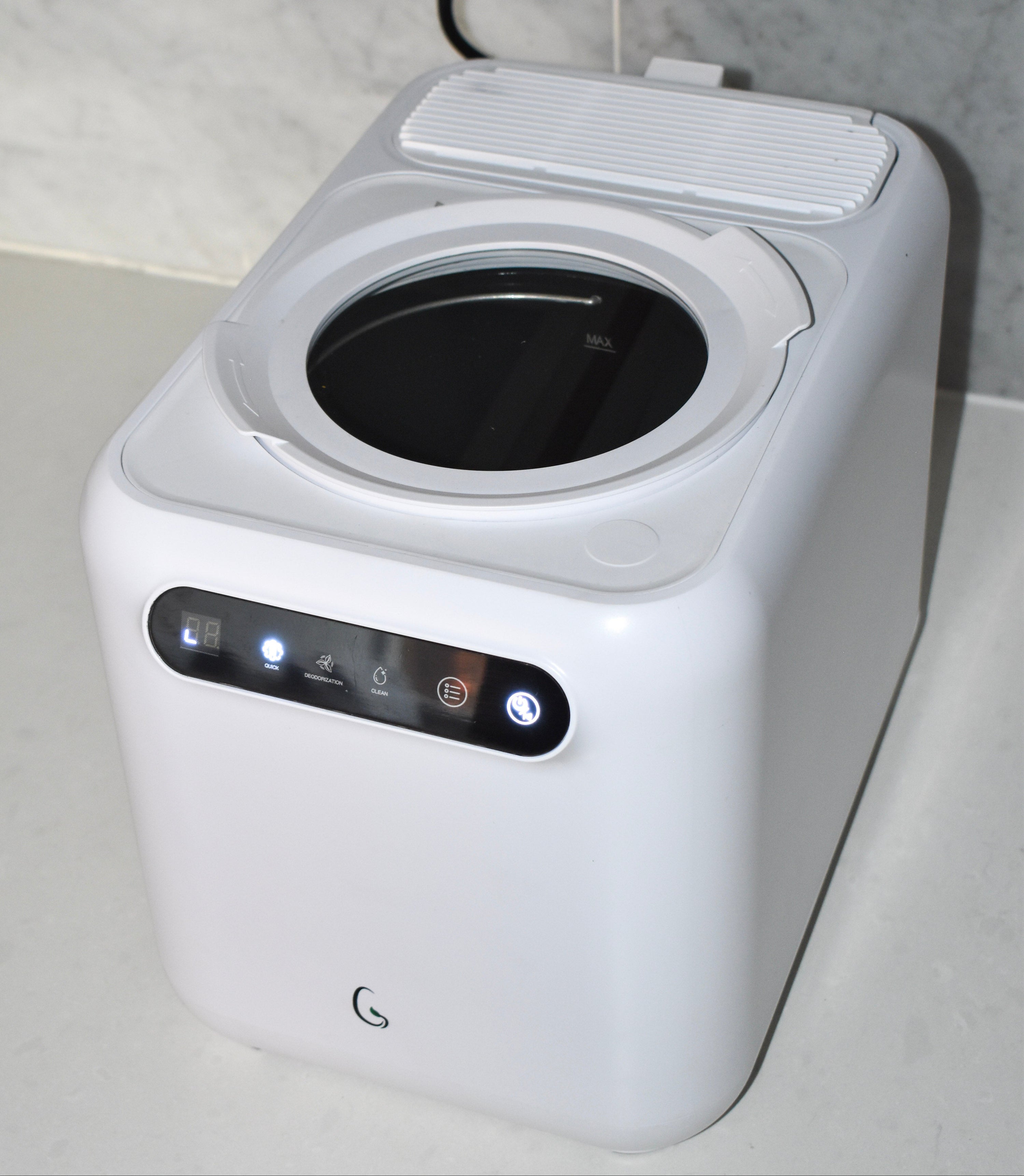 A white countertop appliance with a circular dark opening on top, a front control panel with illuminated buttons, and a power cord plugged in against a tiled wall.